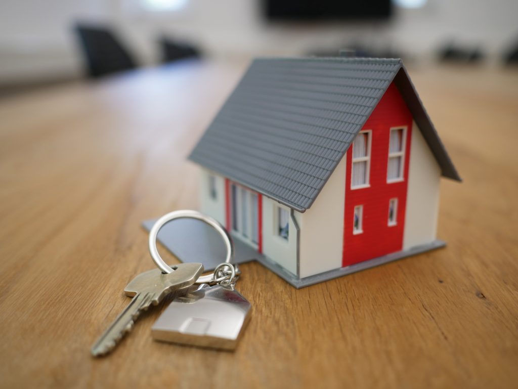 A miniature house standing on a table. The keys are sitting right next to the house.